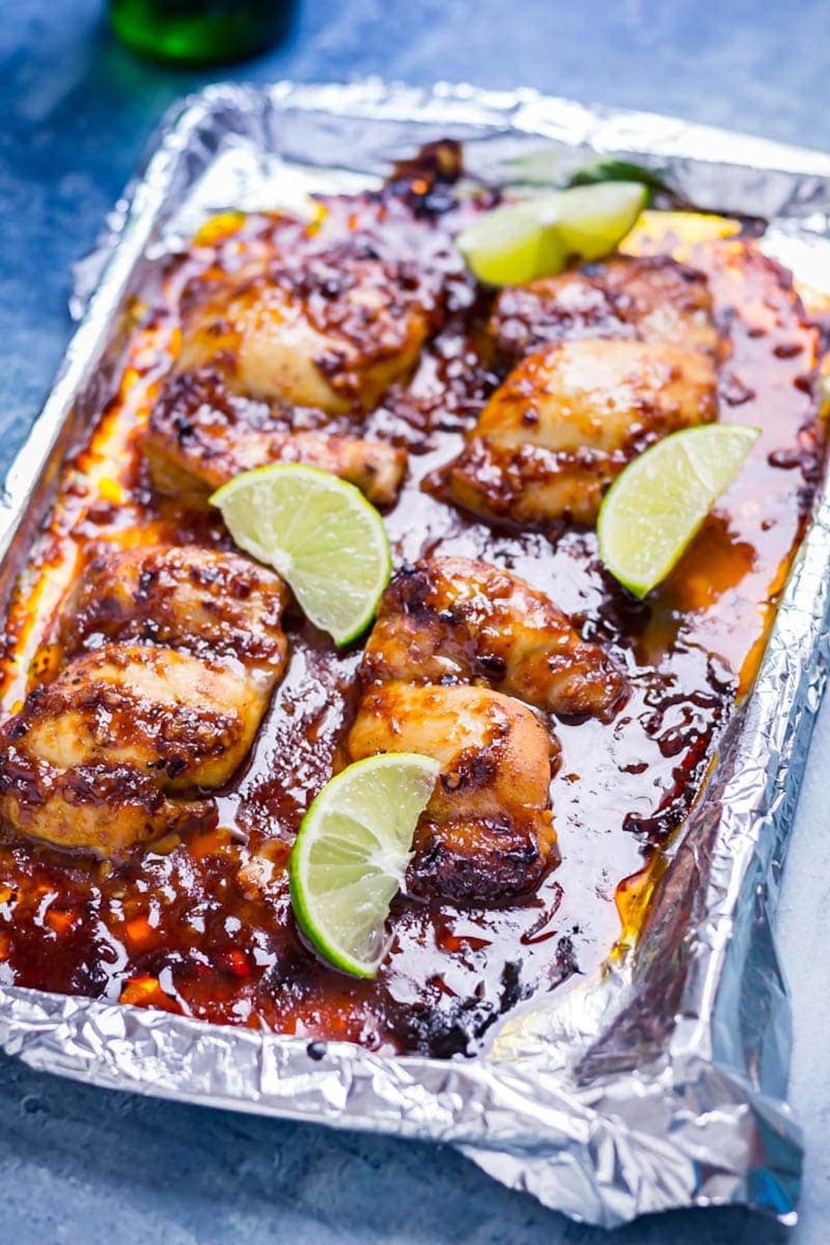 A sheet pan coated in aluminum foil is full of of chicken that's coated in and swimming in a red honey chipotle sauce. Slices of lime are next to each chicken thigh.