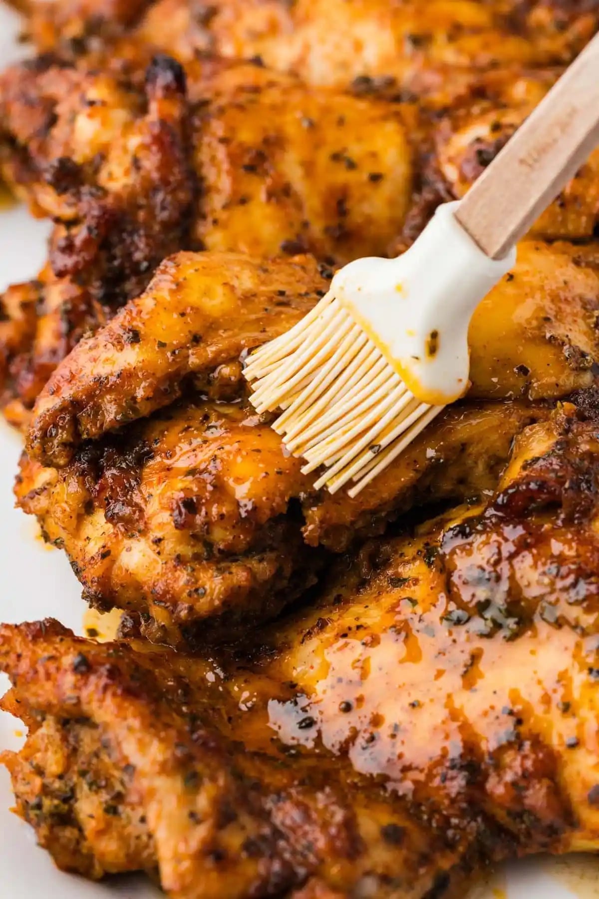 A chicken thigh coated in a reddish yellow marinade is being brushed by a silicone pastry brush. Several other chicken thighs are out of focus in the background.