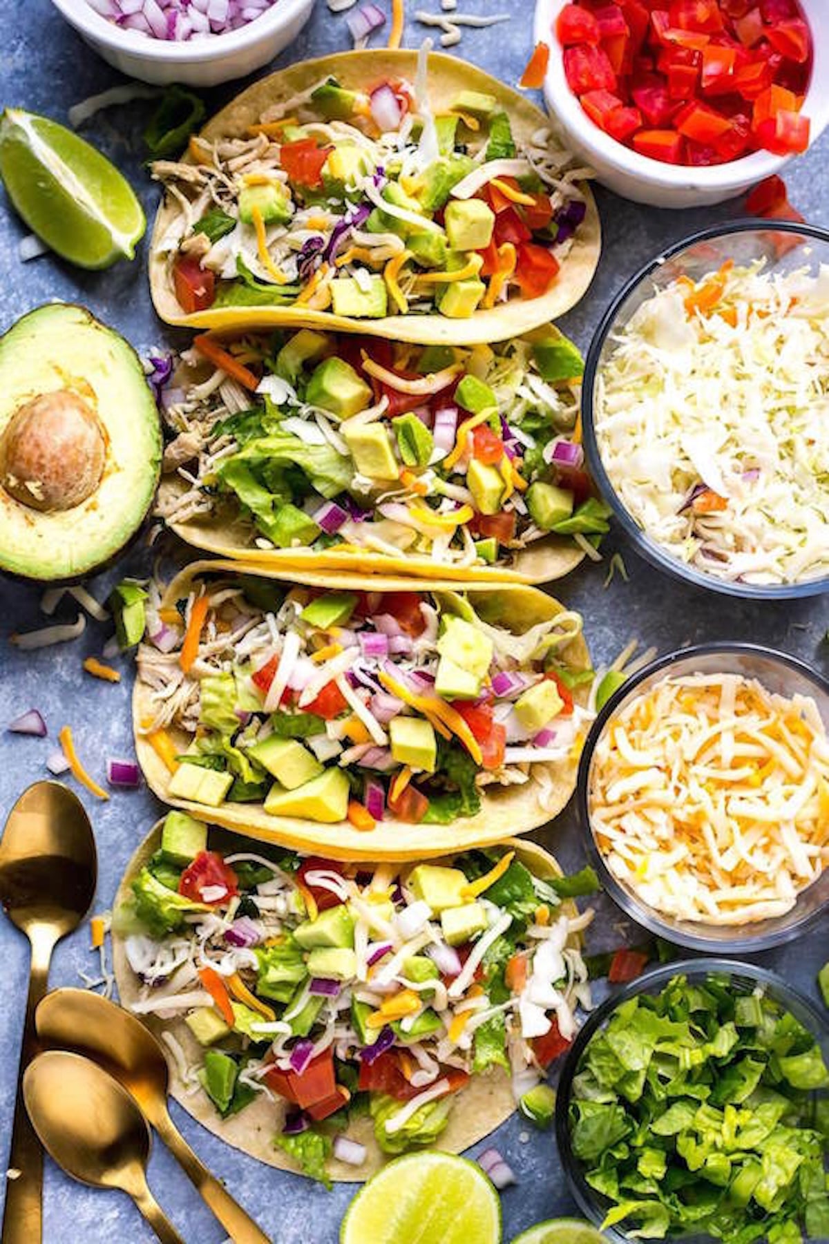 Four corn tortilla tacos generously filled with shredded chicken, chopped avocado, chopped lettuce, and diced red onion sitting next to two bowls of cheese, lime wedges, and a half avocado.