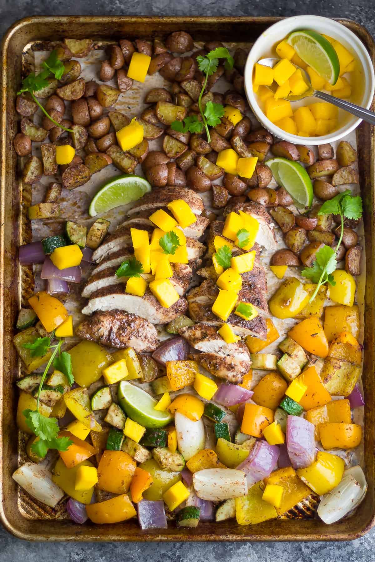 A sheet pan is filled with sliced chicken breast that's coated in a dark marinade, chopped yellow peppers, red onion, and potatoes. A small bowl of diced mango and a lime wedge sits on the side.