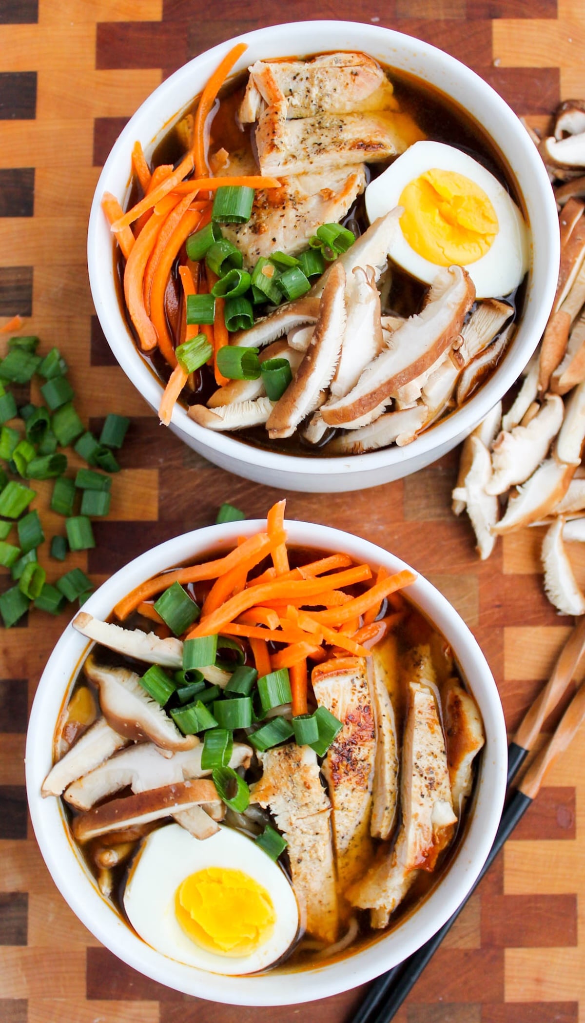 Two white soup bowls each filled with a homemade ramen that's topped with a half hard-boiled eggs, shredded carrots, slide chicken breast, and sliced mushrooms.