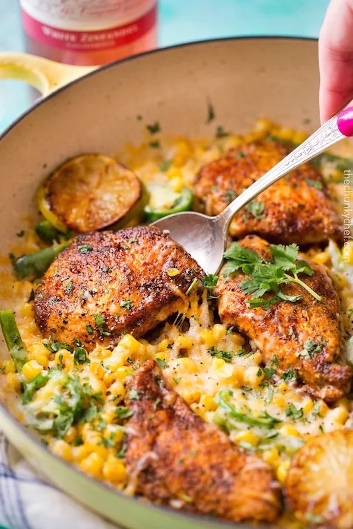 A spoon lifts a golden brown chicken breast out of a skillet that's filled with cheese corn and topped with chopped herbs.