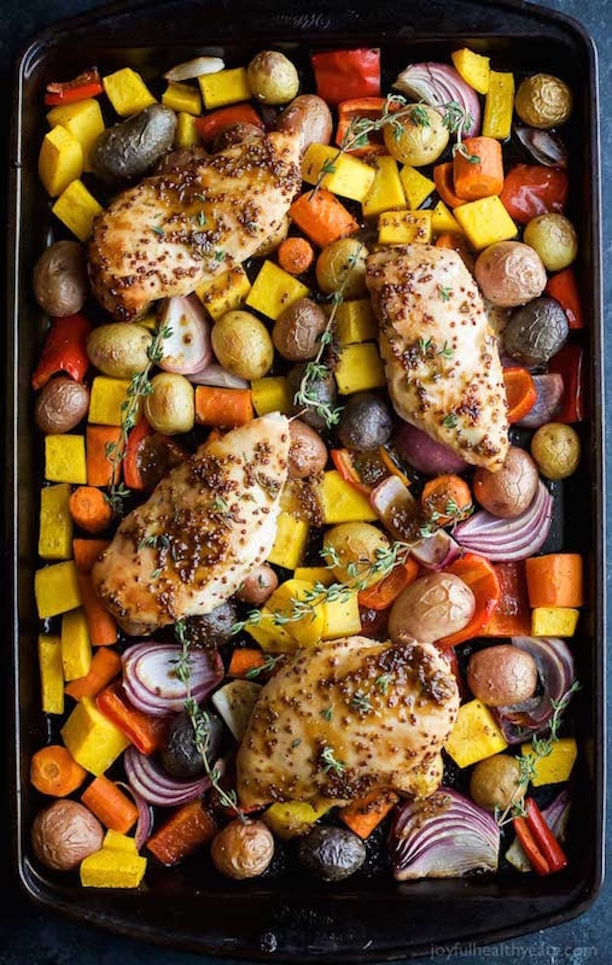 A sheet pan filled with chopped root vegetables, red onion, and seasoned chicken breasts with large sprigs of thyme.