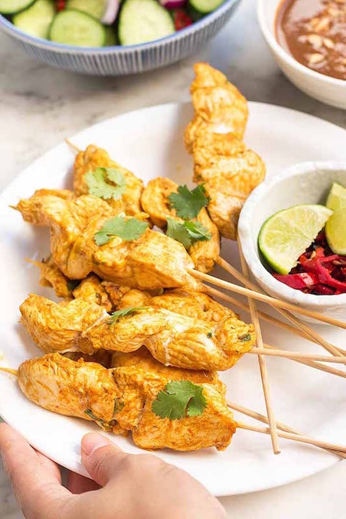 A white serving plate is filled with skewers of chicken that have been marinated in an orange sauce and then cooked. A bowl of limes and spicy peppers is on the side.