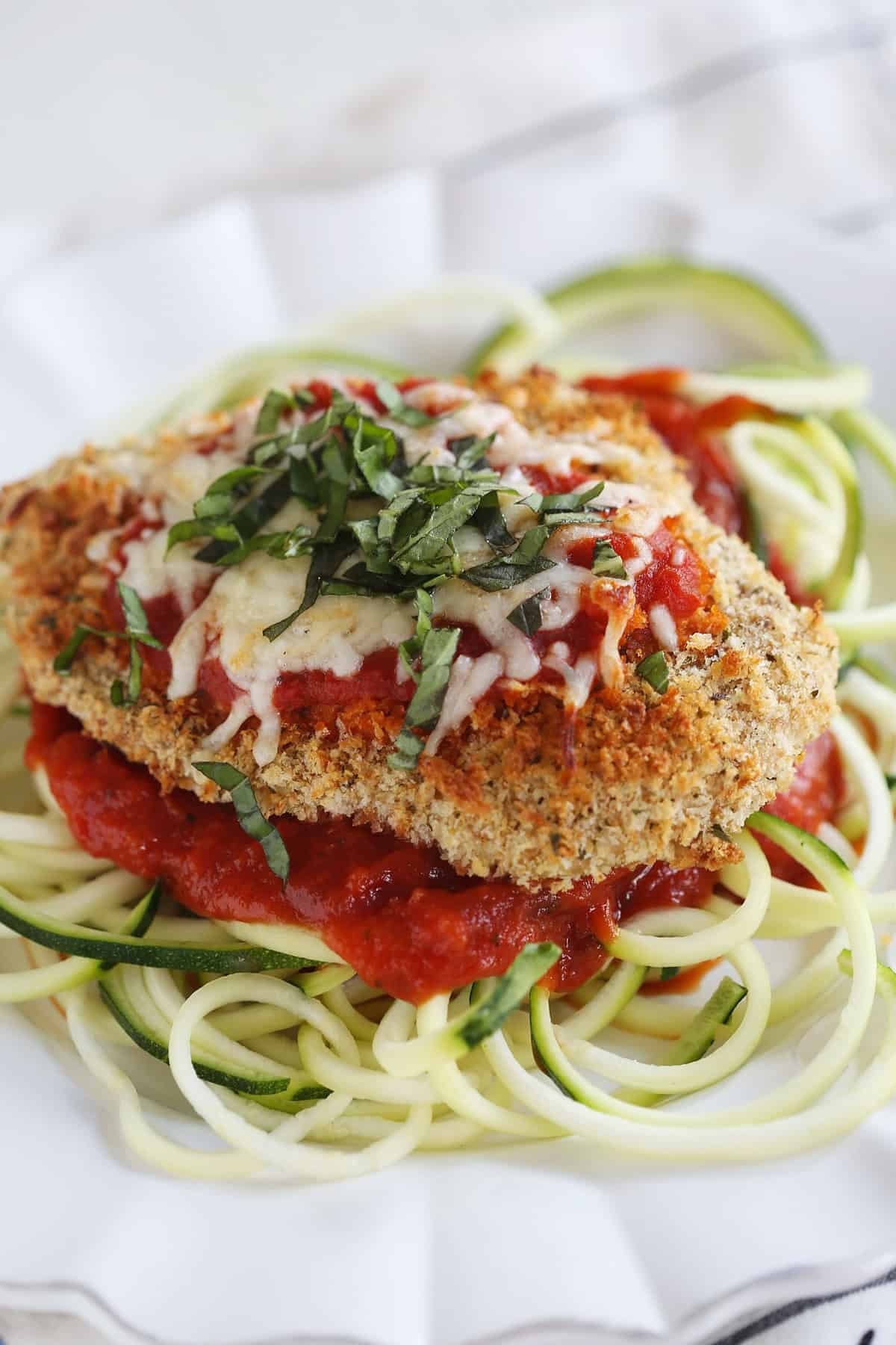 A breaded chicken breast is topped with res sauce and melted white cheese. It sits on a bed of zucchini noodles on a white dinner plate.