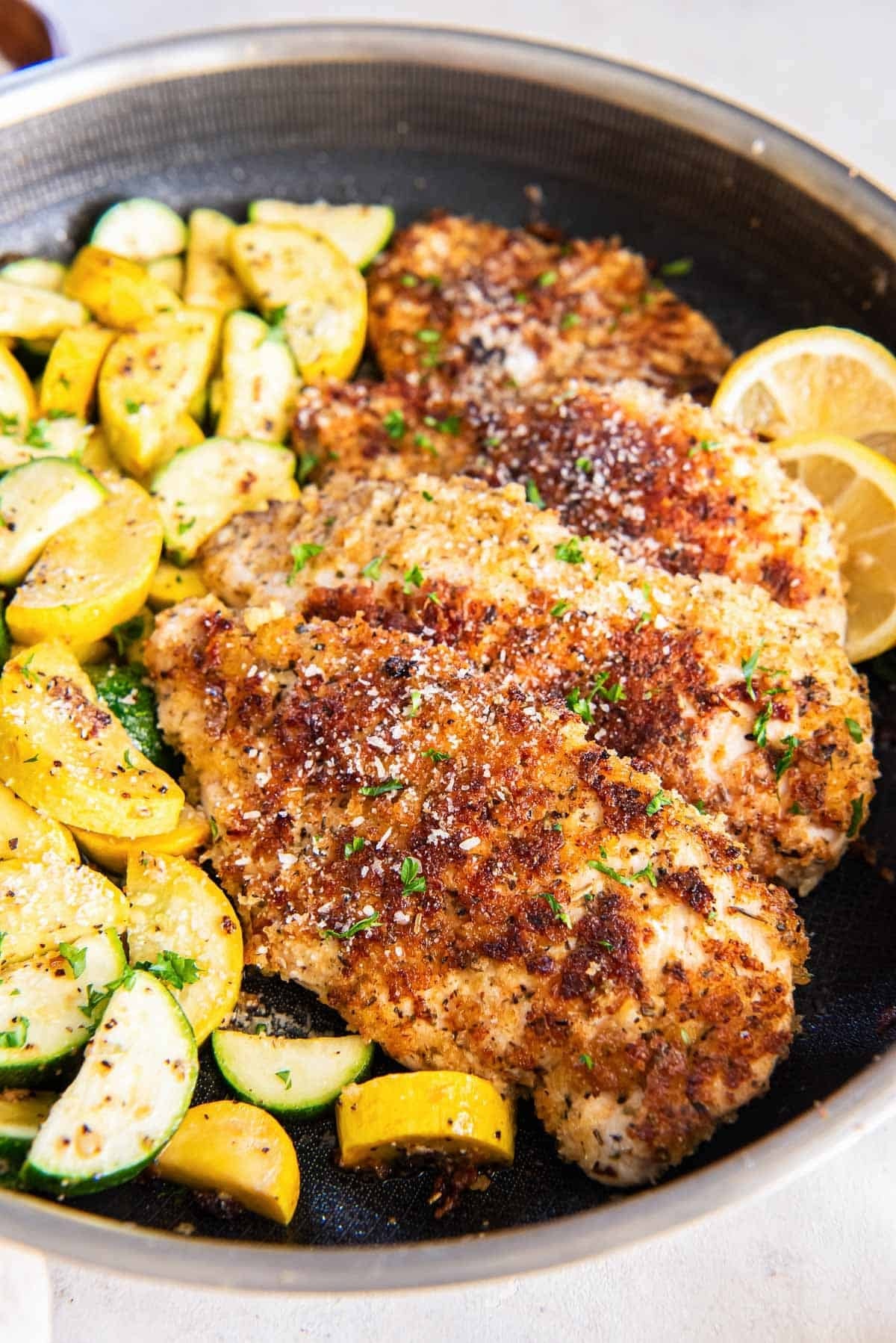 A large skillet has cooked chicken breast that's coated in a golden brown parmesan breading, next to sautéed squash and zucchini moons.