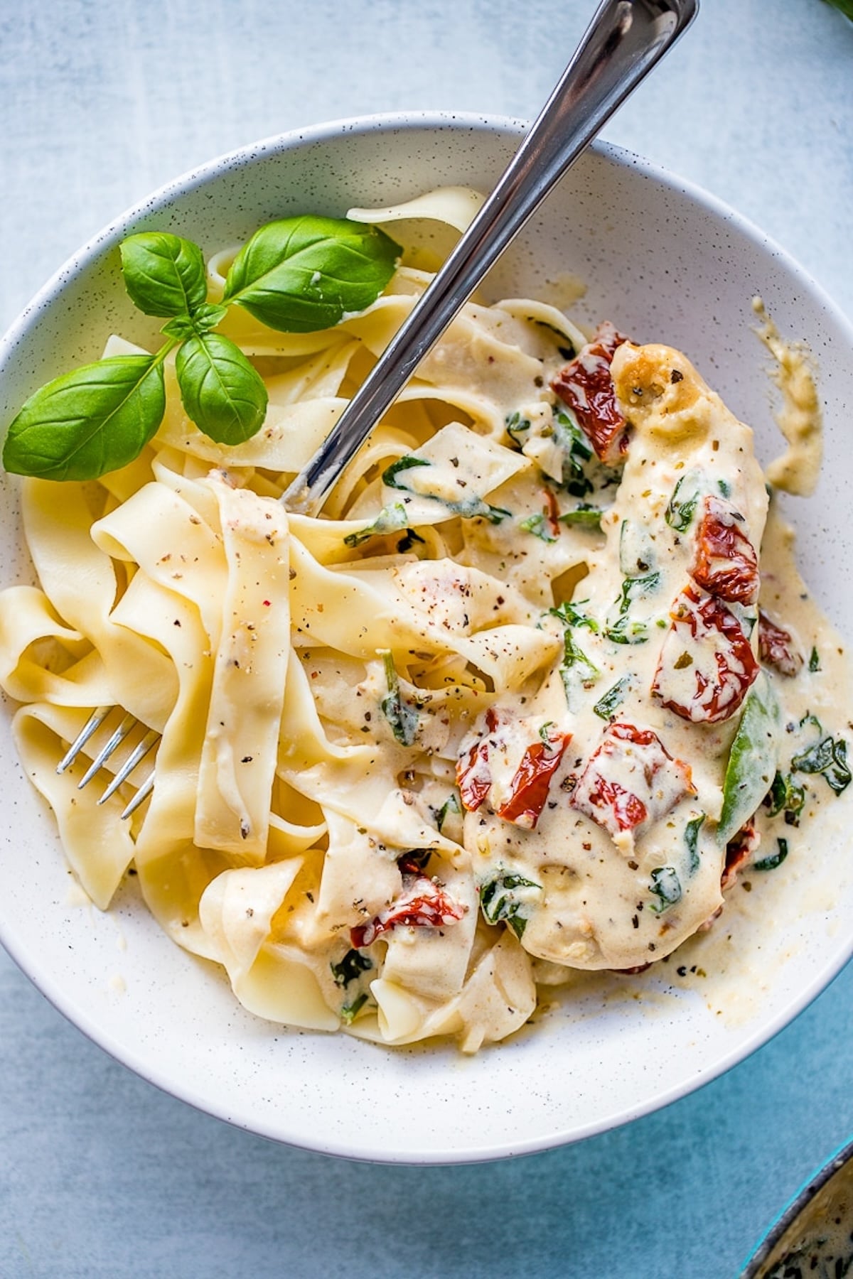 A serving bowl filled with ribbon pasta that's been tossed in a white sun-dried tomato  and spinach sauce. Fresh basil sits on the side.