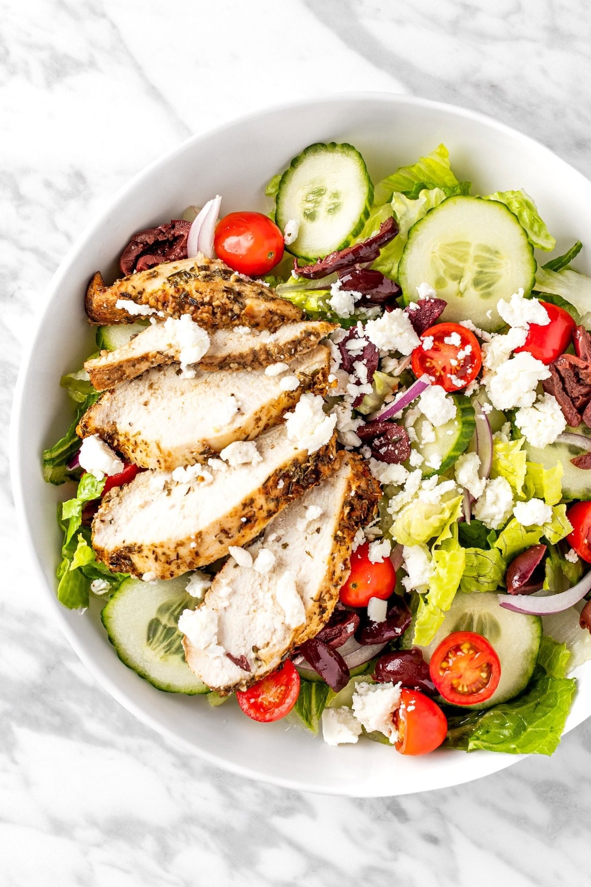 A white salad bowl is filled with roughly chopped lettuce that's topped with slice chicken Brest, sliced cucumber, halved cherry tomatoes, sliced red onion, Kalamata olives, and crumbled feta cheese.