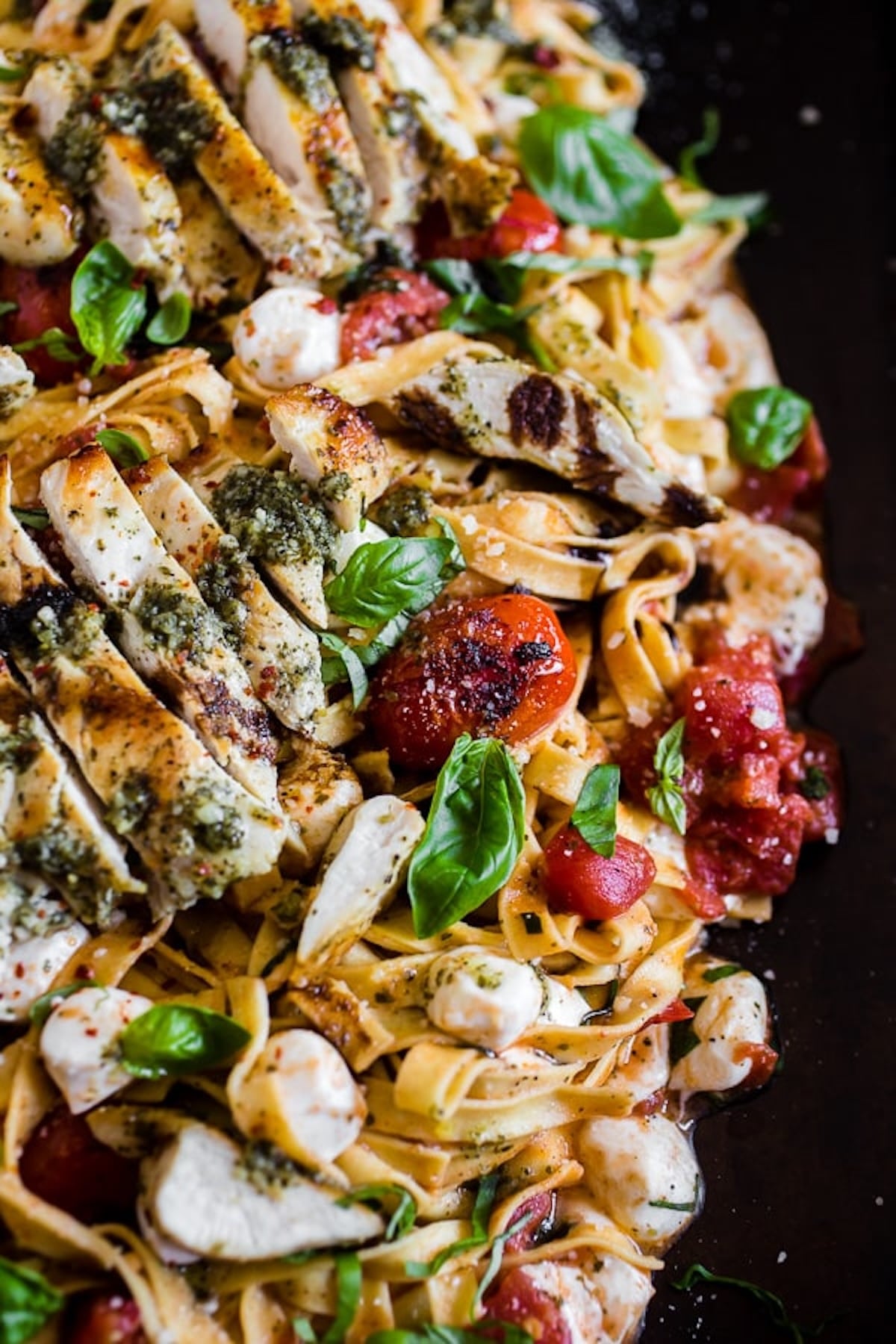 Ribbon pasta topped with seared cherry tomatoes, fresh basil, mozzarella pearls, and sliced chicken breast on top of a black serving plate.
