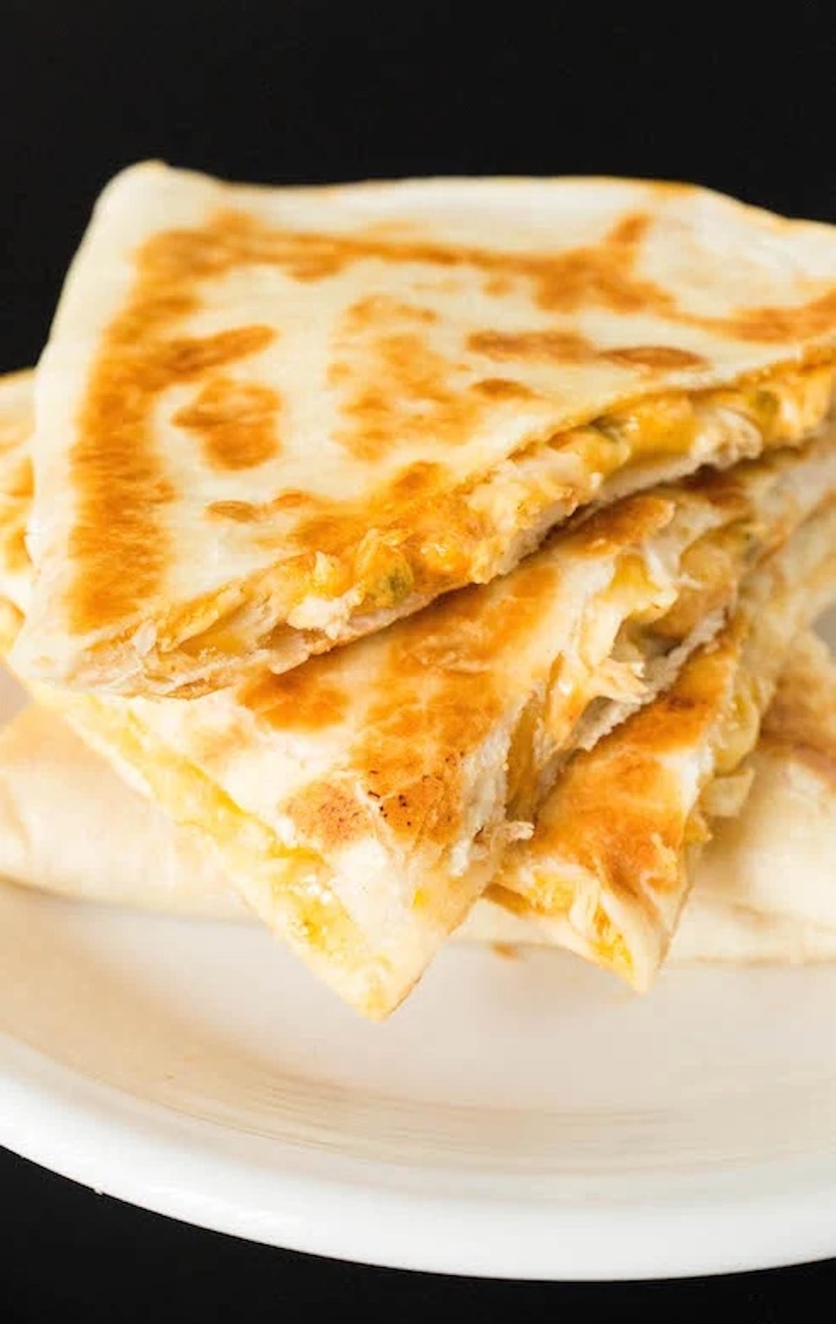Several slices of a chicken quesadilla have been stacked on top of one another on a small serving plate. You can visibly see pieces of chicken breast, melted cheese, sauce, and diced jalapeños.