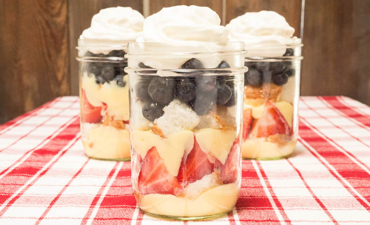 Three mason jars filled with layers of angel food cake, strawberries, blueberries, pudding and whipped cream.