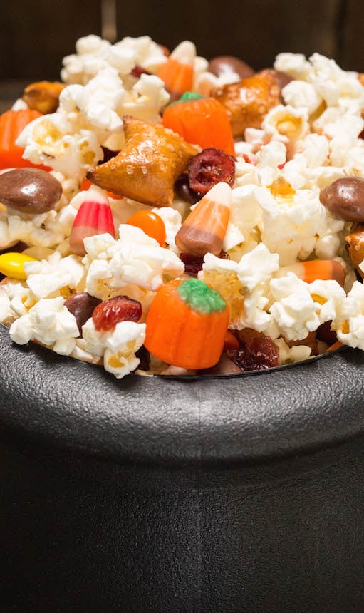 A plastic cauldron filled with Halloween Snack Mix with popcorn, candy corn, pumpkin candies, cranberries, peanut butter filled pretzels, and milk duds.