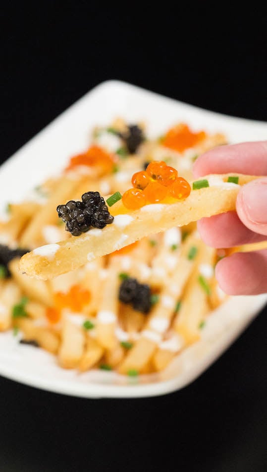 A hand holds up a French fry that's topped with two different kinds of caviar and chives.