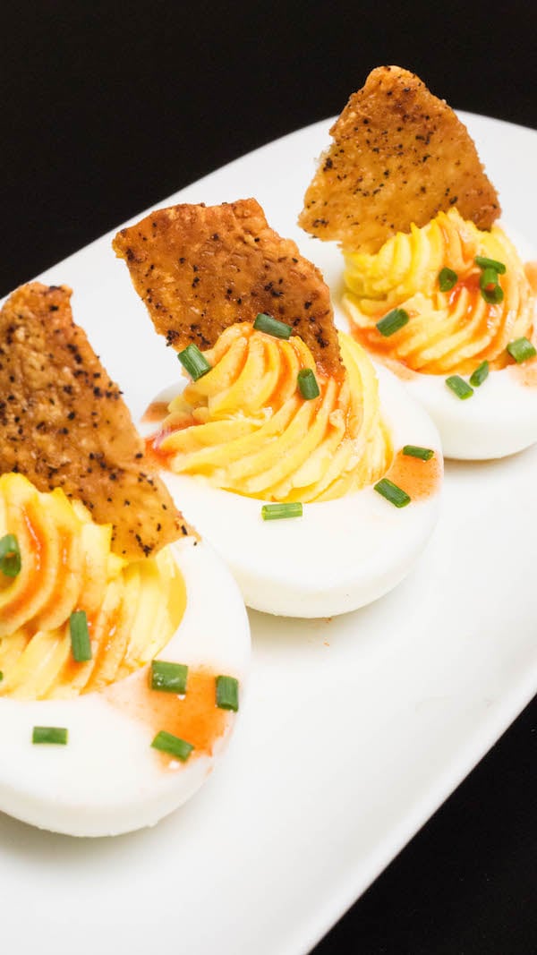 A white serving plate with Deviled Eggs topped with Fried Chicken Skin & Hot Sauce.