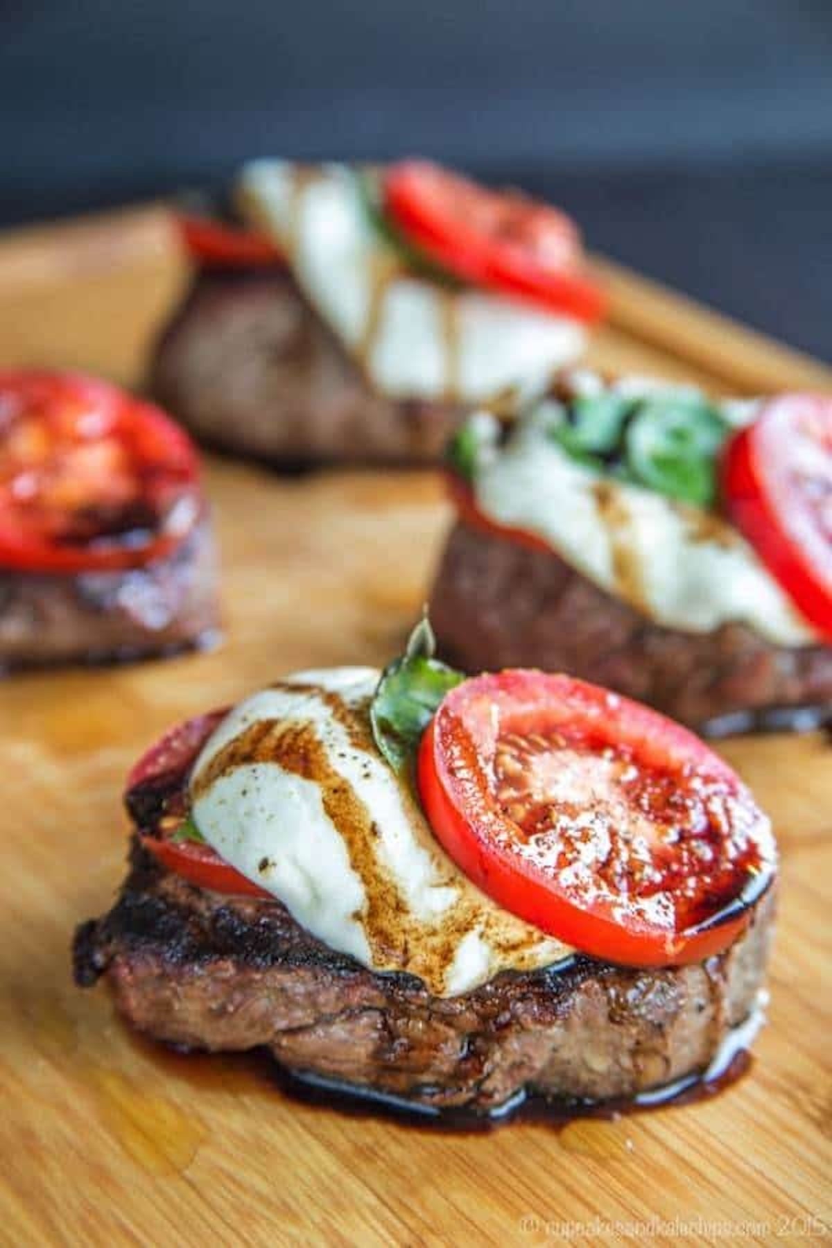 A wooden board has several grilled filet mignon steaks, each topped with mozzarella, a tomato slice, fresh basil, and a balsamic drizzle.