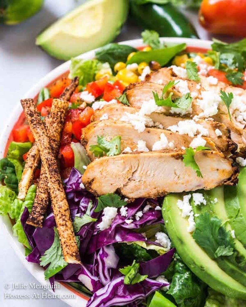 A white bowl is filled with a salad made of mixed greens and purple cabbage, sliced grilled chicken, sliced avocado, tomatoes, and crumbled white cheese and cilantro.