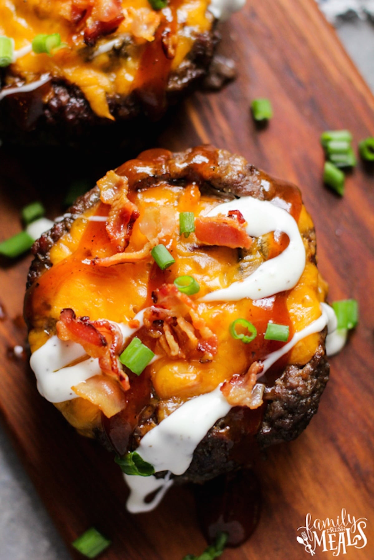 A cooked burger patty has been hollowed out in the middle and filled with melted cheese, chopped bacon, and sliced green onions then drizzled with ranch.