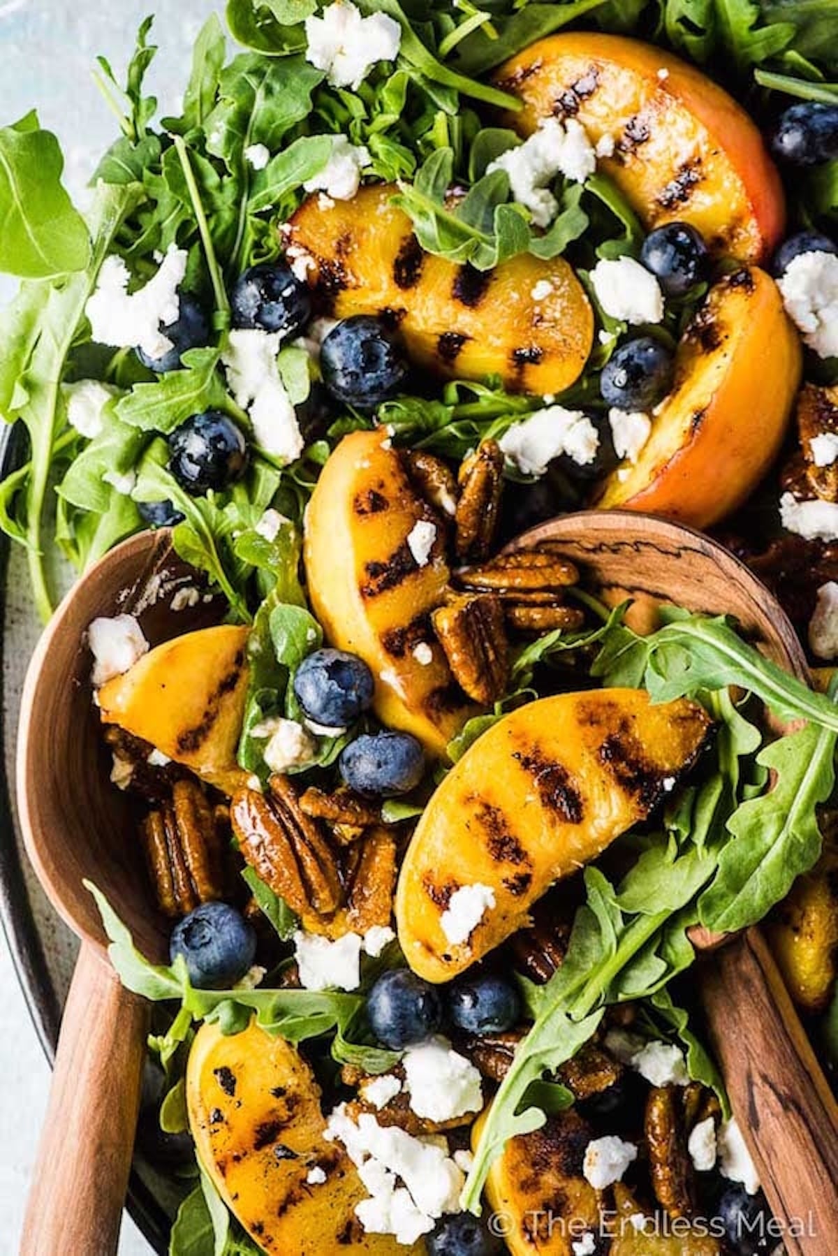 A salad with arugula as the base and topped with grilled peach slices, blueberries, crumbled white cheese, and pecans.
