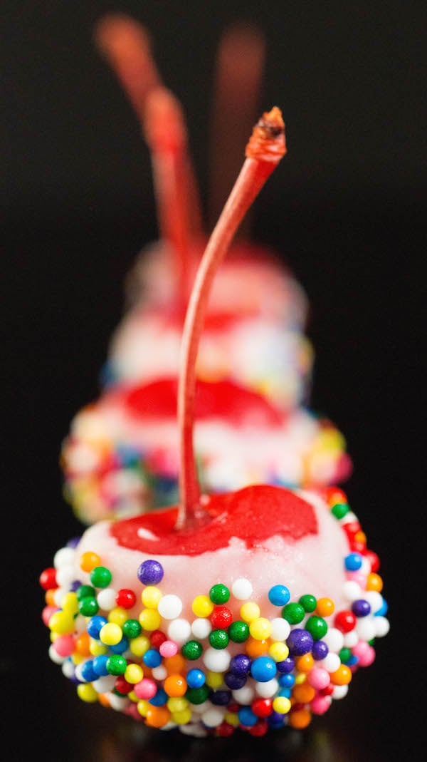 Birthday Cake Cherry Bombs (maraschino cherries soaked in cake vodka then dipped in white chocolate and rainbow sprinkles) on a black background .