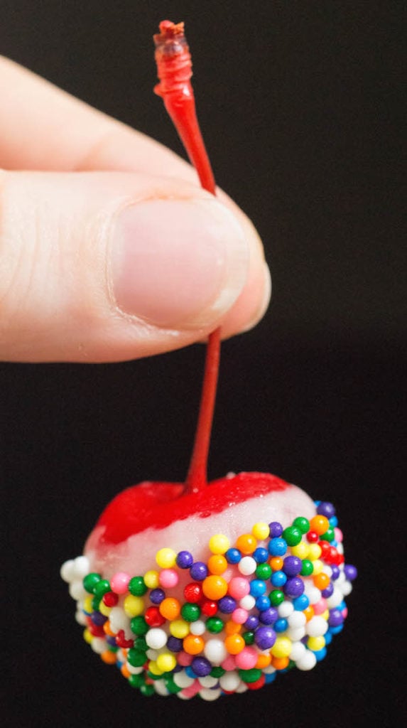 A hand holding up a maraschino cherry that's been soaked in cake vodka then dipped in white chocolate and sprinkles.