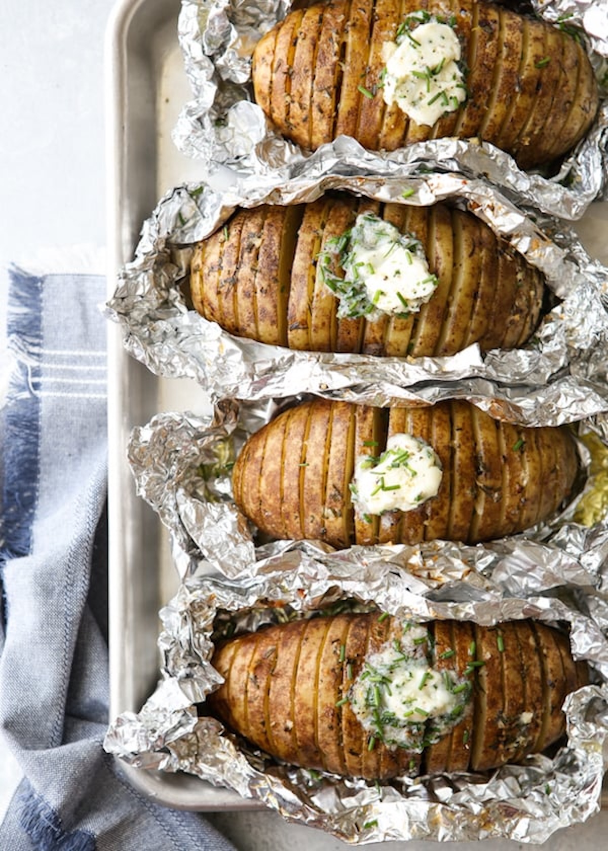 Four hasselback style sliced potatoes are topped with a chive butter, wrapped in foil, and placed on a sheet pan.
