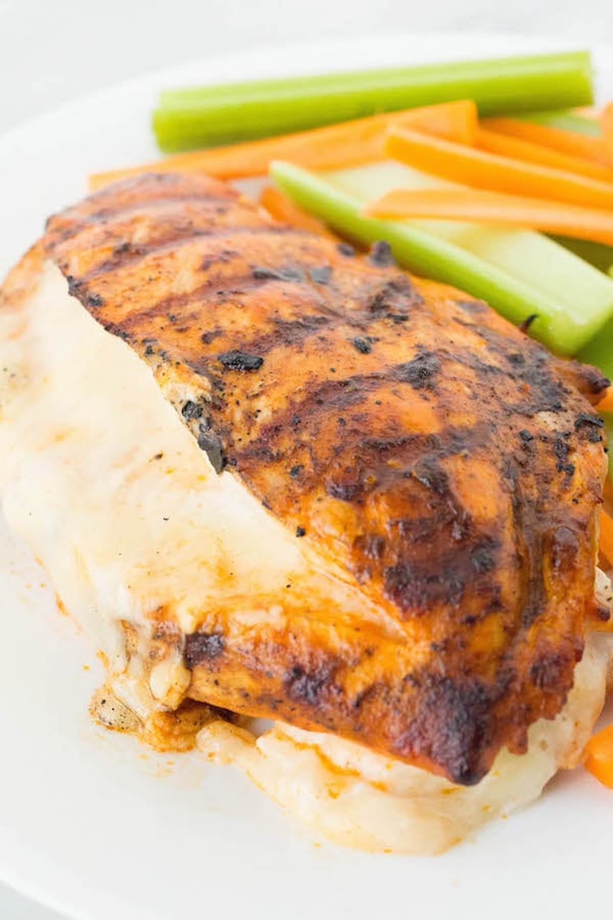 A grilled chicken breast brushed with buffalo sauce has been filleted and is oozing melted white cheese from the middle. Celery sticks and carrot sticks are out of focus in the background.
