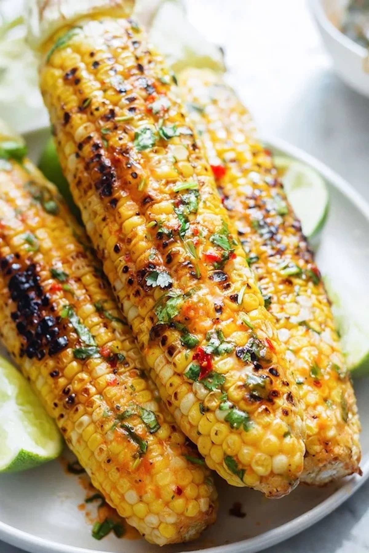 Grilled corn on the cob topped with chopped herbs and hot peppers are stacked on top of one another. A sliced lime is out of focus in the background.