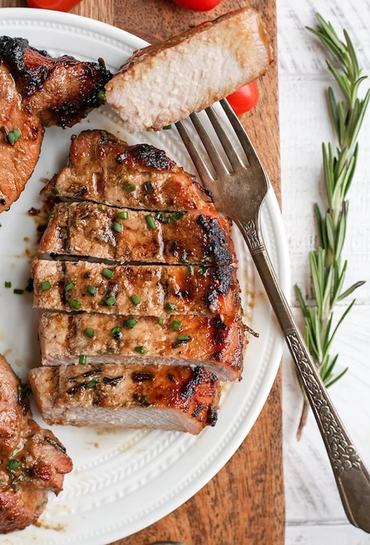 A grilled pork chop has been cut into thick slices on a dinner plate. Fresh herbs are off to the side.