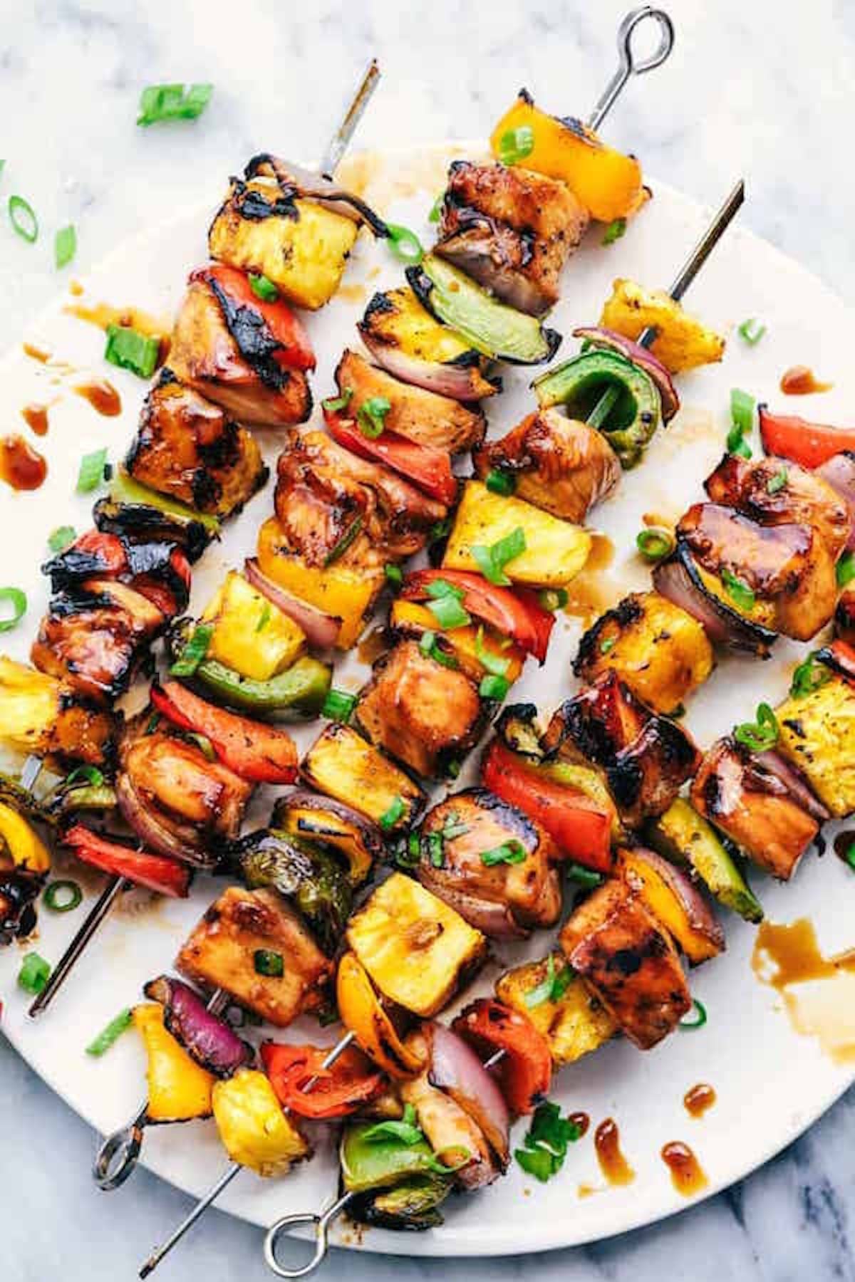 A white dinner plate is filled with skewers that feature grilled chubed chicken, pineapple, red peppers, green peppers and red onion all brushed with a teriyaki glaze.