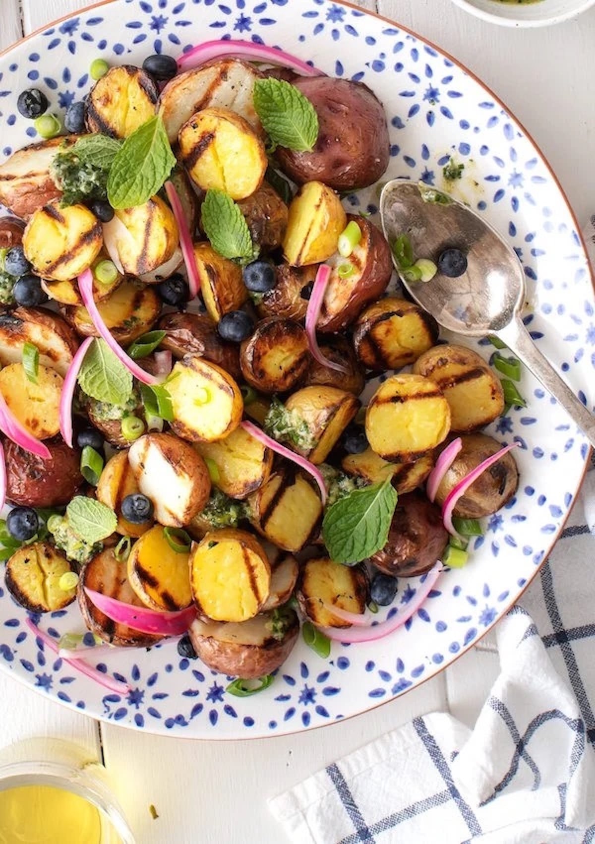 A serving bowl is filled with small , halved, grilled potatoes, red onion, sliced green onion, blueberries, and mint.