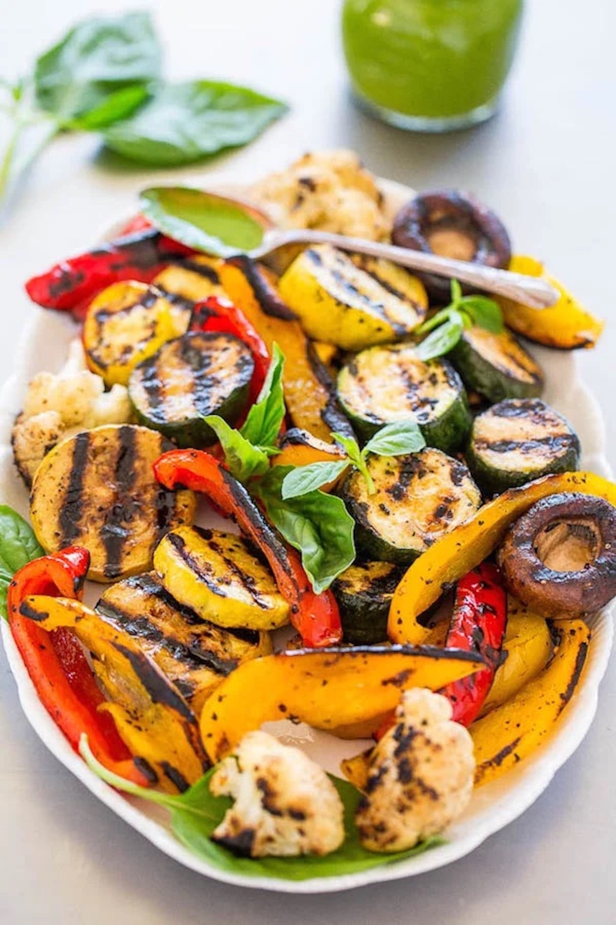 A large white serving bowl filled with sliced vegetables and baby Bella mushrooms that have visible grill marks. A green sauce in a jar is out of focus in the background.