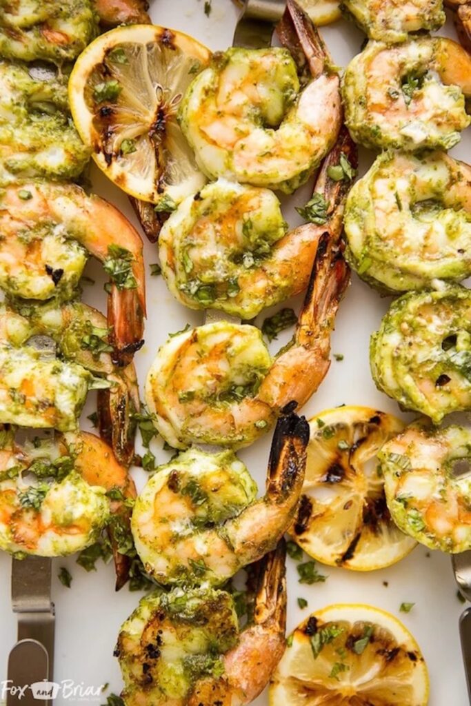 Dozens of grilled shrimp coated in a lemon pesto sauce on skewers. Grilled slices of lemon lay around them.