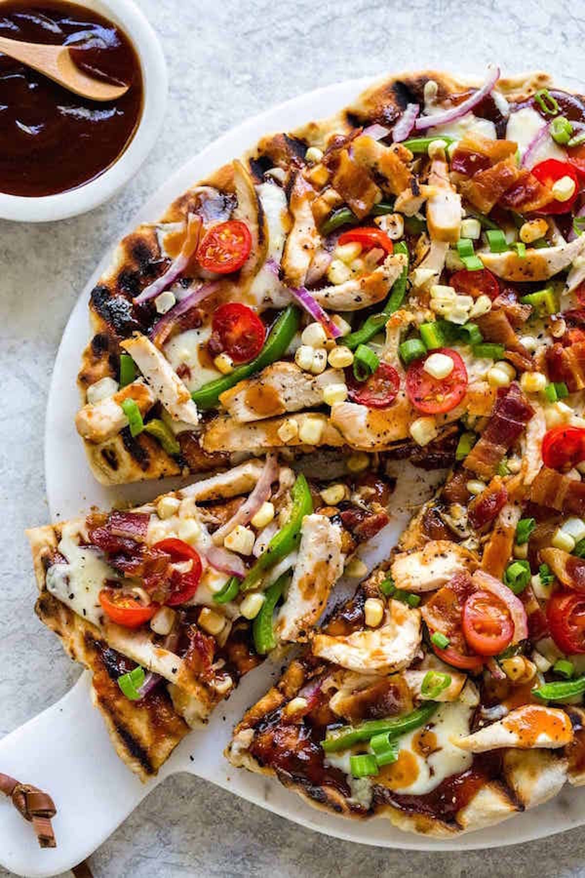 A thin crust pizza is loaded with cheese and bbq sauce and toppings like sliced peppers, red onion, cherry tomatoes, corn kernels, and chicken.