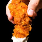 A close-up of a hand holding a homemade buffalo chicken tender that's been dipped in ranch sauce against a black background