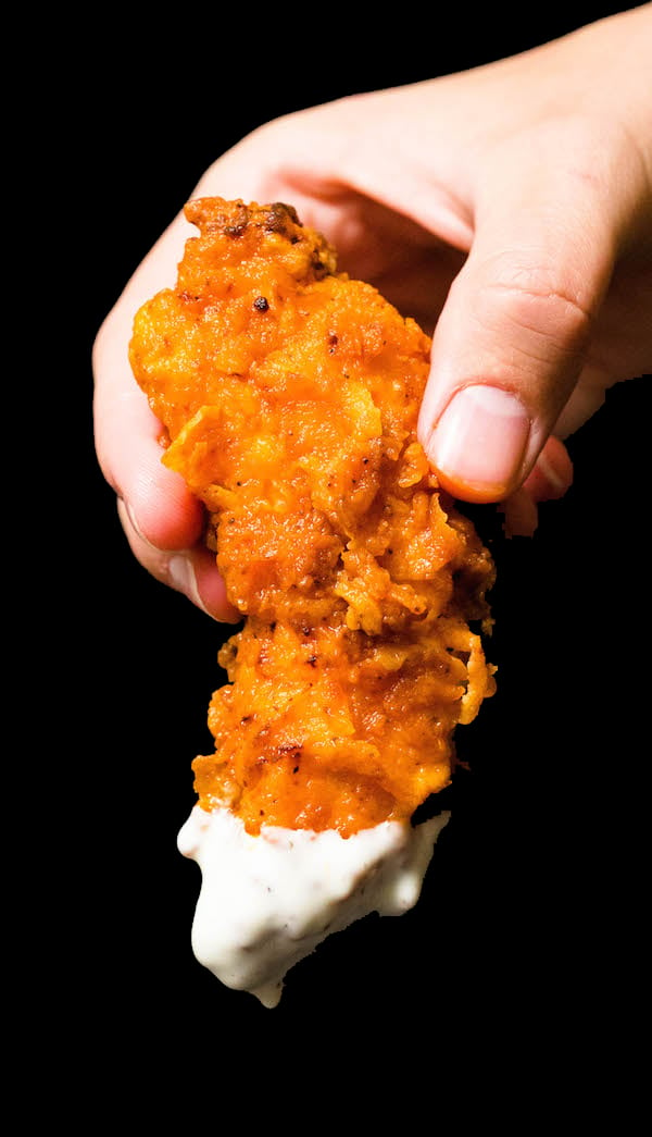 A close-up of a hand holding a homemade buffalo chicken tender that's been dipped in ranch sauce against a black background