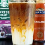A pint glass is lined with caramel sauce and filled 1/3 of the way with milk and the rest with espresso. A bag of coffee beans and a bottle of caramel sauce are out of focus in the background.