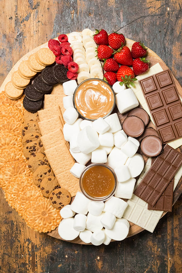 Overhead view of large round wood board topped with marshmallows, caramel sauce, peanut butter, chocolates, and various cookies to make s'mores.