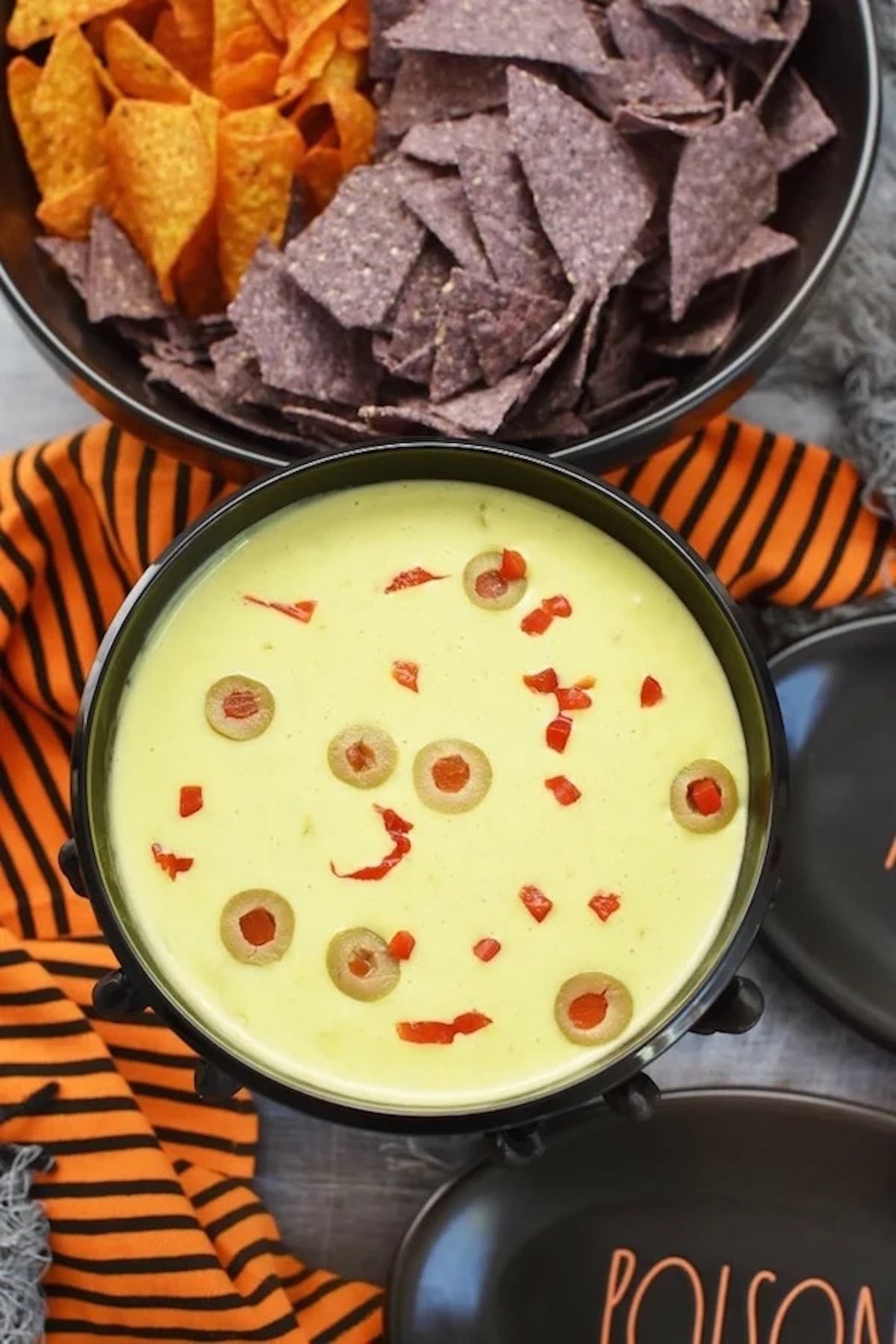 A green dip is garnished with floating sliced green pimento stuffed olives. Orange and purple tortilla chips are out of focus in the background.