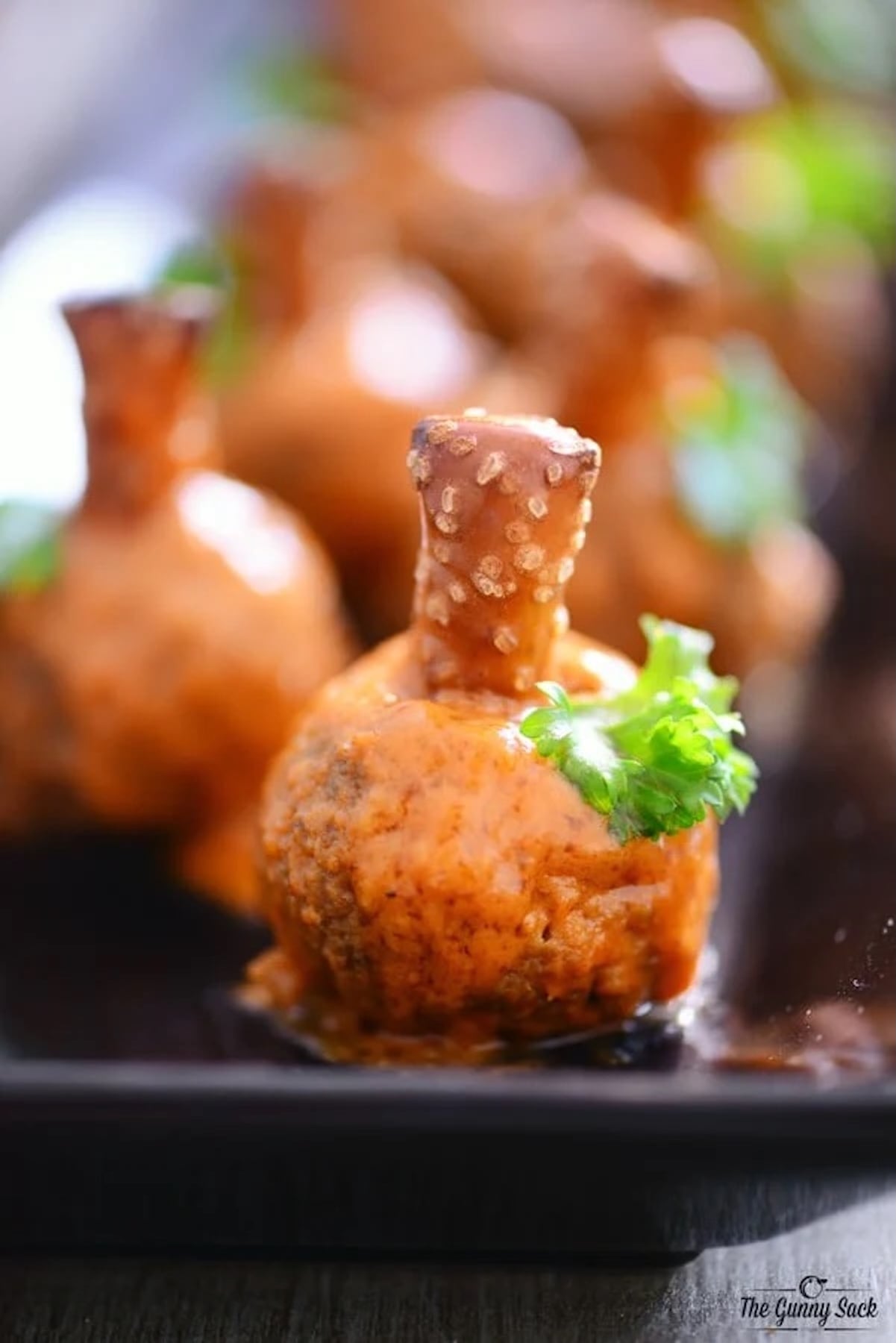 A meatball resembling a pumpkin is covered in orange sauce and a pretzel rod sticking out of the top to look like a stem. A piece of cilantro is placed on the side to look like a leaf.