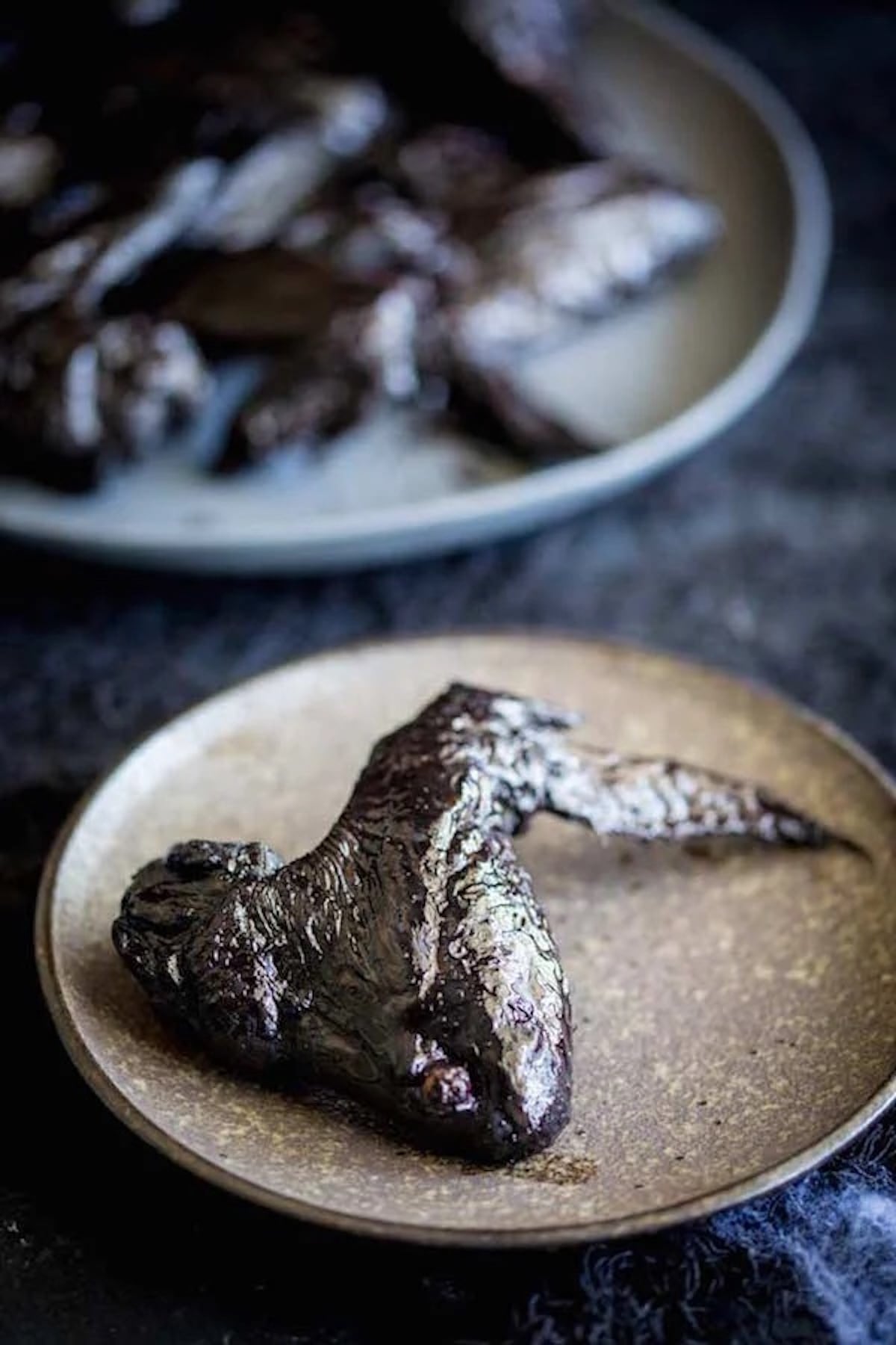 A whole chicken wing that has been dyed black to look like a bat wings on a small gray plate. More wings are out of focus in the background.