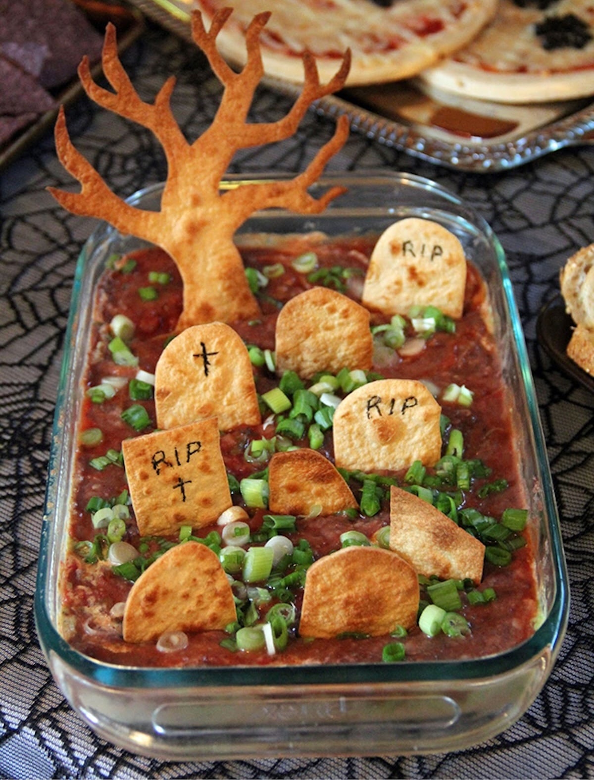 A glass 16x9 dish is filled with a red sauce and baked tortilla chips that have been cut into the shapes of headstones. "RIP" is written on a few.