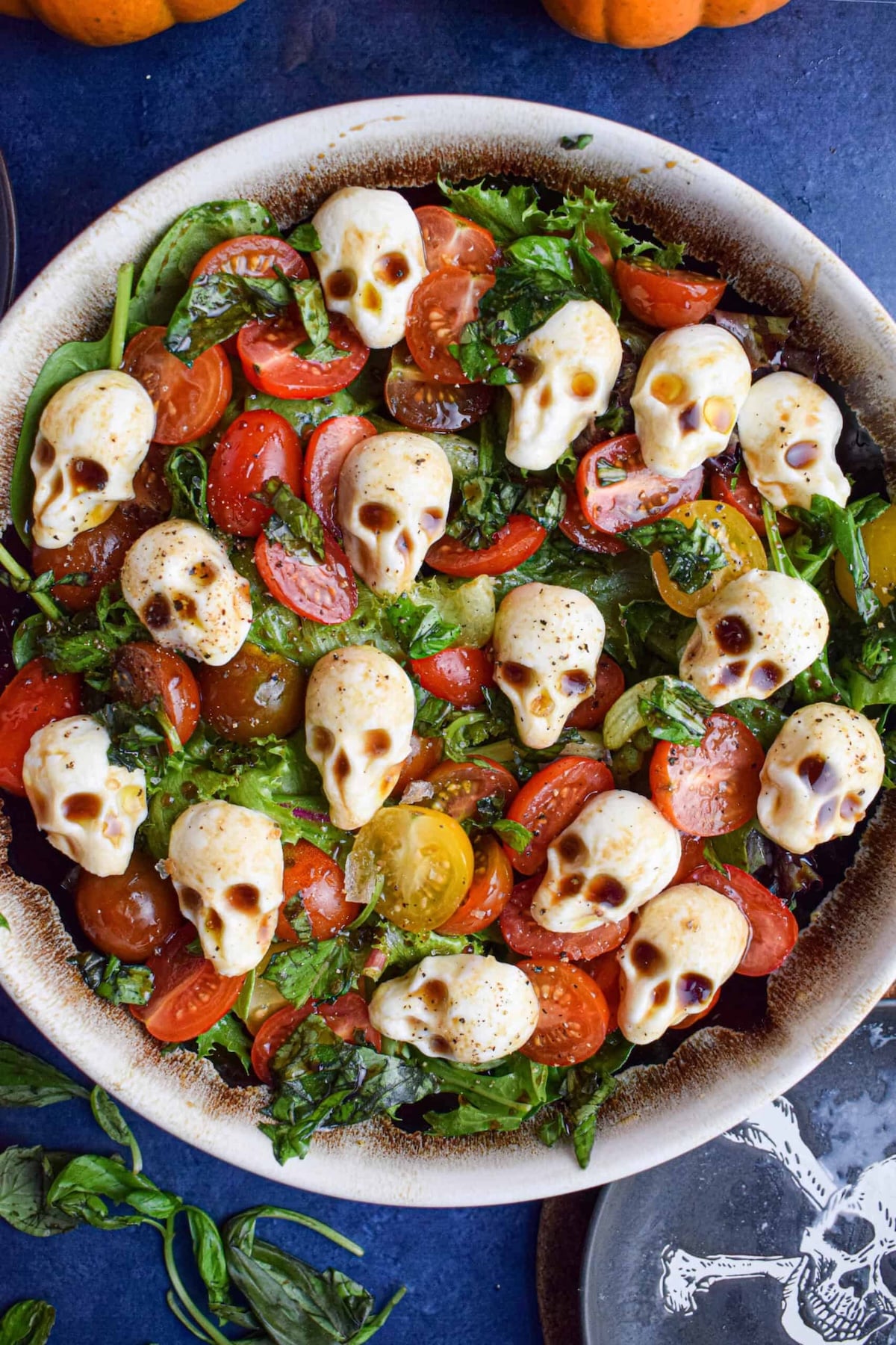 A bowl is filled with a salad that's topped with halved cherry tomatoes and mozzarella pearls that are shaped like skulls.