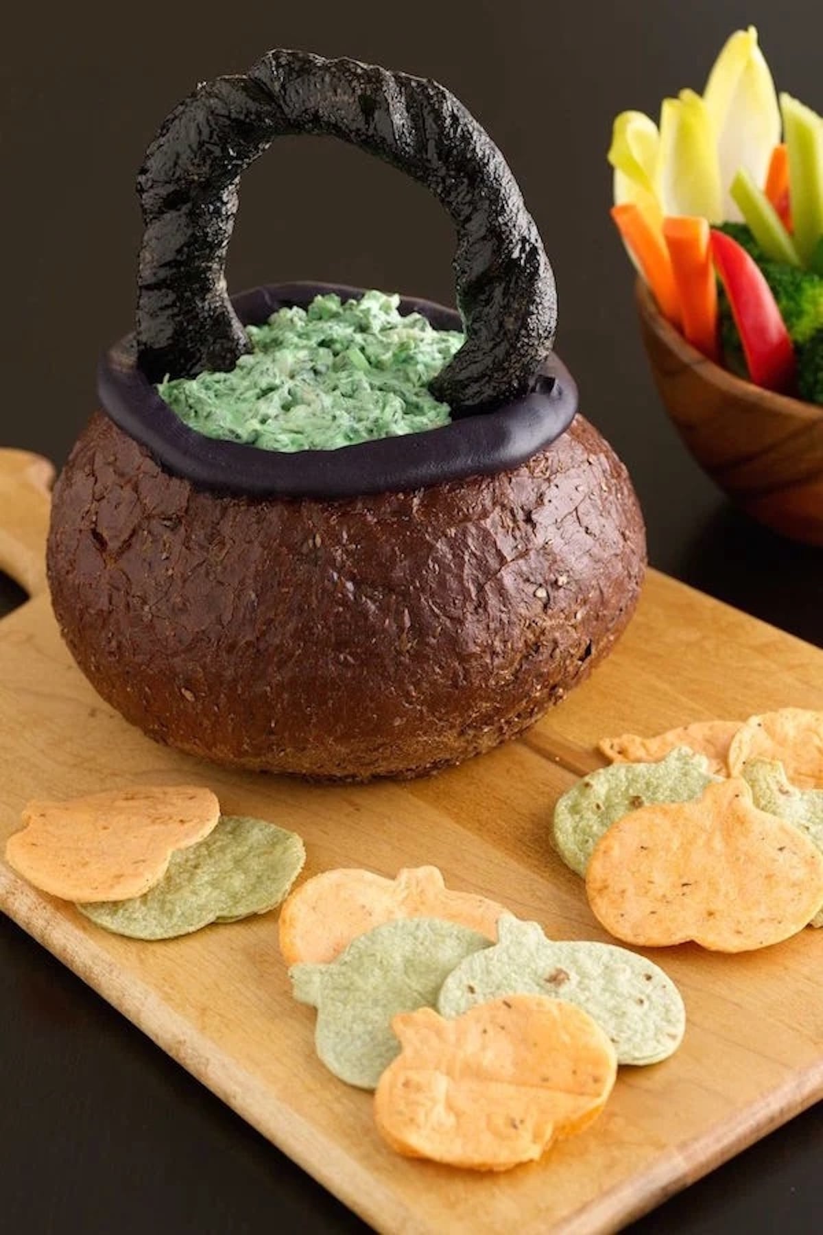 Brown bread is shaped into a cauldron and filled with a green, chunky spinach dip. Pumpkin tortilla chips are arranged around the cauldron.