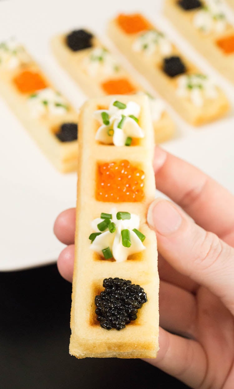 A hand holds up a Caviar Waffle Stick. A platter with the rest of them is out of focus in the background.