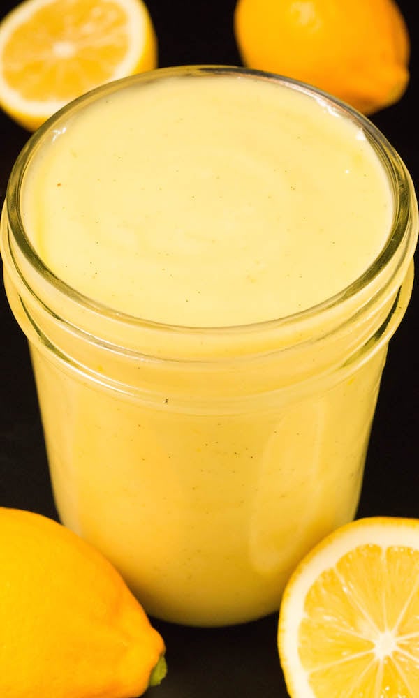 A small glass jar filled with Vanilla Bean Meyer Lemon Curd surrounded by cut lemons.