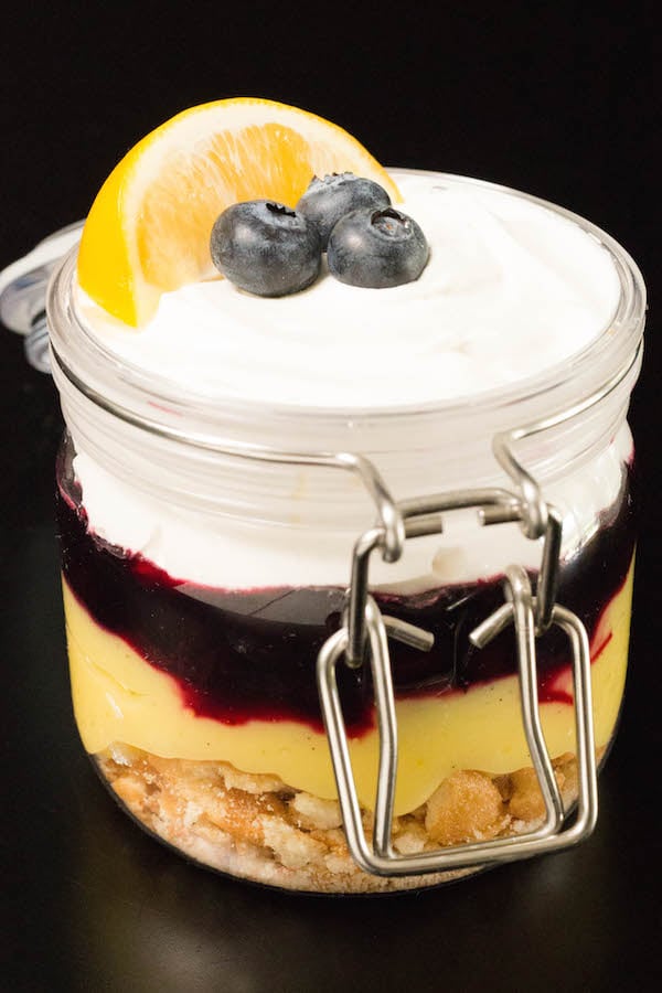 A meyer lemon curd parfait in a jar on a black background. It's garnished with a lemon slice and three fresh blueberries.