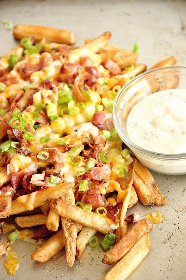 A pile of fries covered in cheese, bacon, and green onions sits on a sheet pan next to a small bowl of ranch dipping sauce