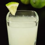 A single lime drop shot in a square glass on a black background with two limes.