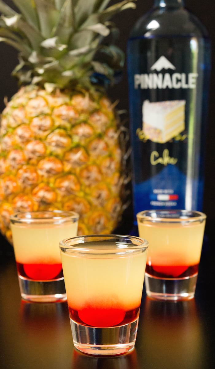 Three layered pineapple upside down cake shots in front of a vodka bottle and a pineapple