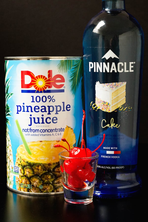 A large can of pineapple juice, a bottle of cake vodka, and a shot of maraschino cherries on a black background.